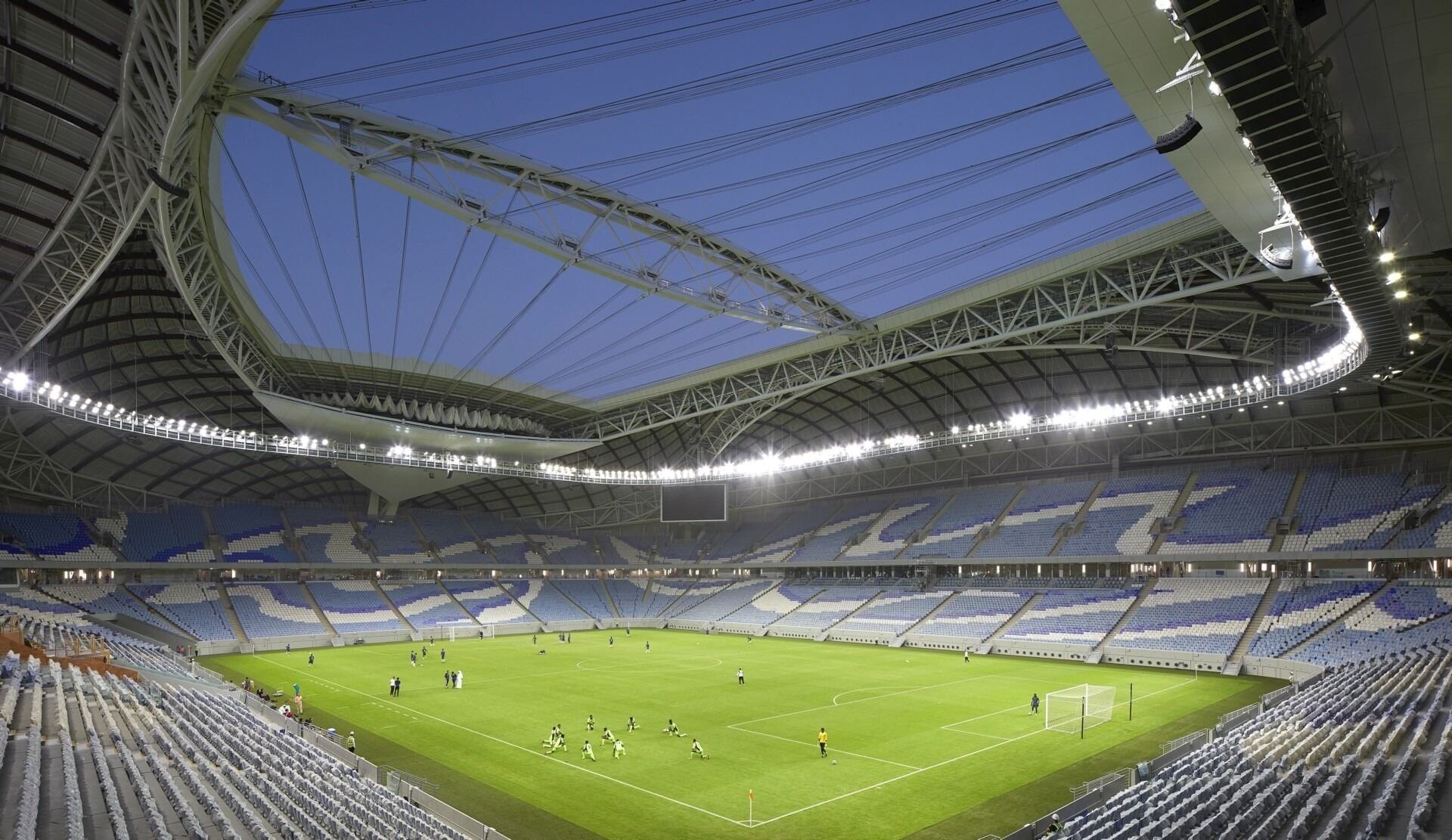 Interior of Al Wakrah Stadium at dusk, showing the steel roof structure and cable-supported closure system above the pitch.