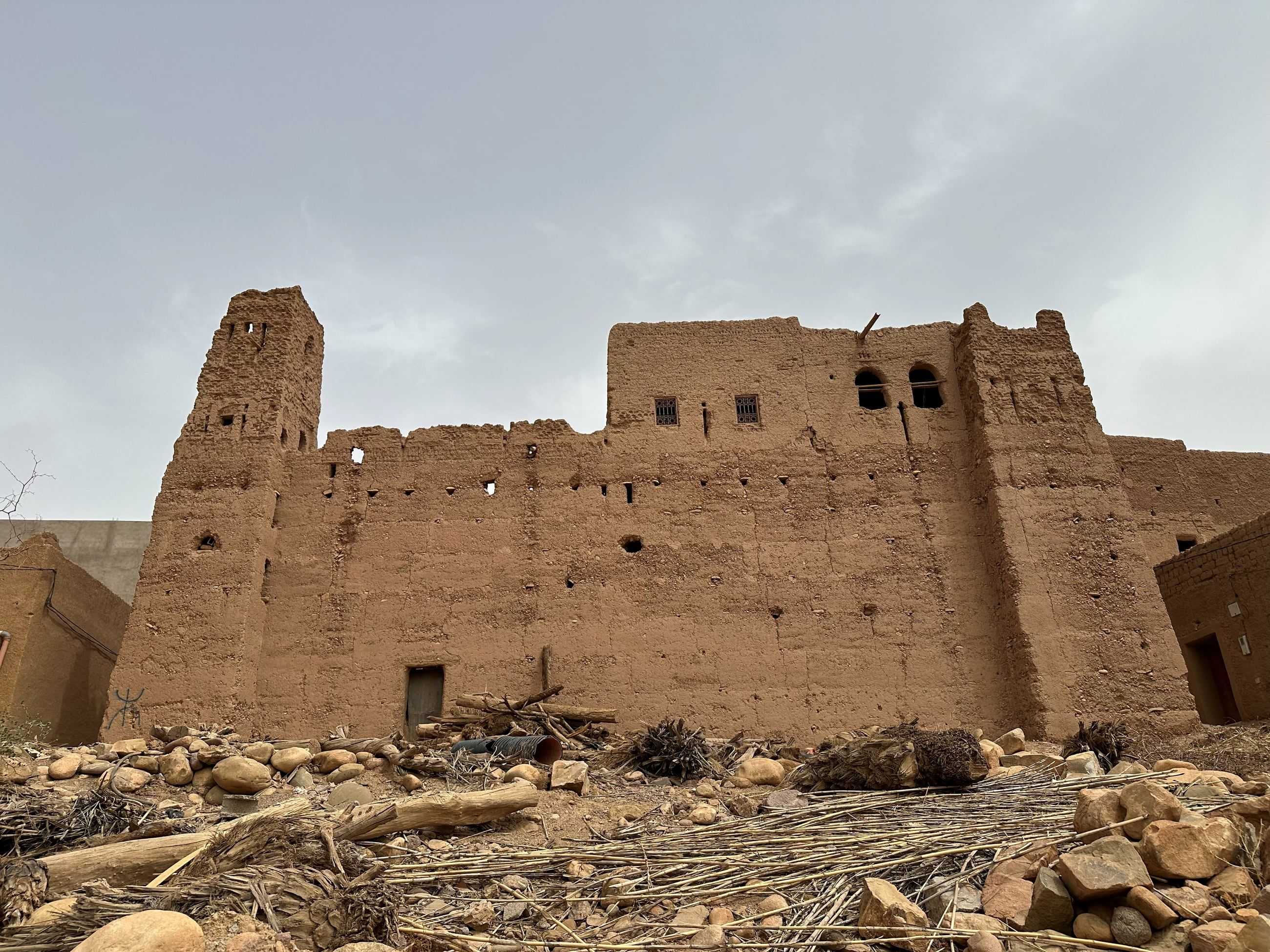 A traditional earthen ksar in Tazarine, Morocco: a long rammed-earth facade with a corner tower and rows of putlog holes, seen across fallen stone and palm debris.